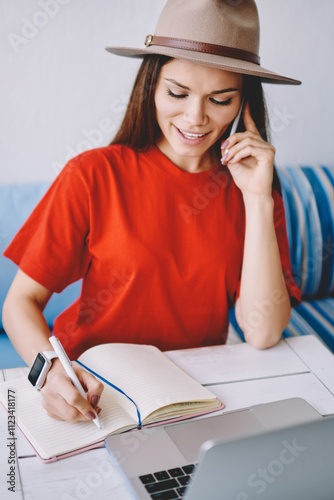 Positive female student calling to customer service for consultancy and writing main theses in textbook, successful woman checking account balance via telephone conversation and notes information photo