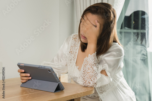 An Asian woman in a white satin nightgown experiences eye pain and fatigue as she reads her work schedule on a tablet, causing her to stay up past her bedtime. photo