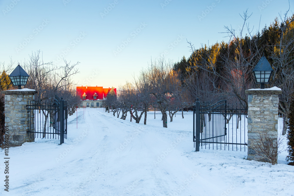 Naklejka premium Traditional stone house with steep red metal roof seen at the end of an alley covered in fresh snow during a winter morning, Saint-Nicolas, Quebec, Canada