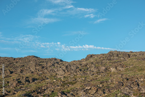 Nature layered background with sunlit grassy rocky hill under clouds in blue sky. Minimalist view to stone hill in bright sun in changeable weather. Natural backdrop of rock mountain pass in sunny day