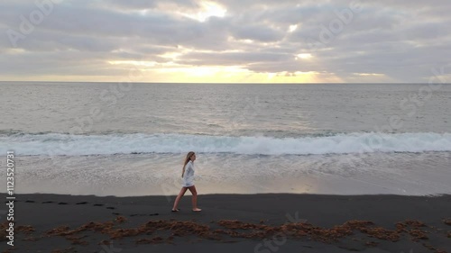 Woman walks along black sand beach at sunset near ocean waves