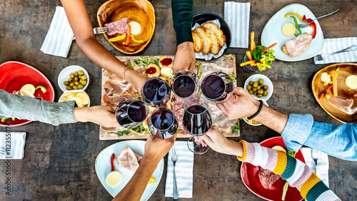 Friends cheering red wine glasses on table covered with delicious food at at farm house vineyard - Top view of people having dinner party at bar restaurant - Food and beverage lifestyle concept