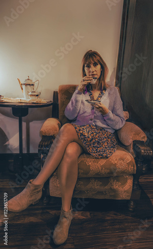 Woman sitting on a sofa holds a cup in her hand and a plate of a tea set she is drinking in her hand