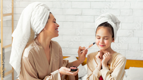 Happy young mom and her teen daughter having beauty day, applying makeup, relaxing and having fun on bed at home. Teen girl and her mother in bath towels using cosmetics, spending time together photo