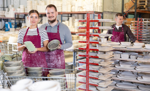 Pair of positive professional artisans, young woman and bearded man in stained burgundy aprons standing in well-lit pottery studio, holding glazed ceramic pieces with pride photo