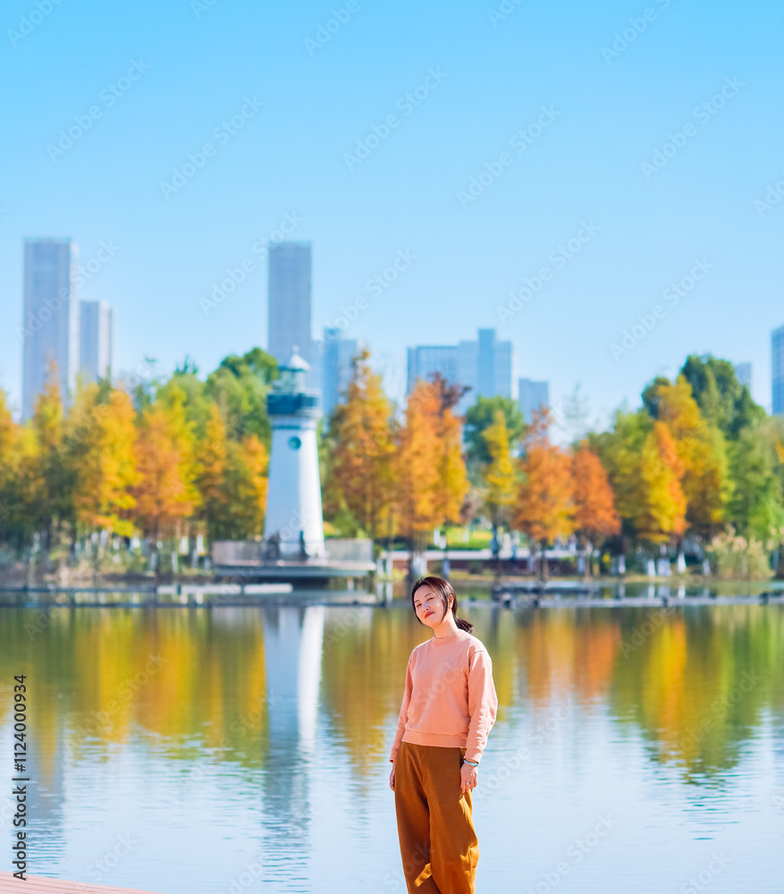 Fototapeta premium One Asian woman enjoying beautiful landscape of golden autumn season at Yinzhou Park, with lighthouse and urban CBD skyline in Ningbo, Zhejiang, China