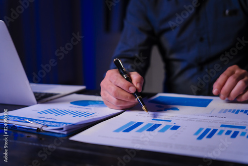 A man works late at night at his desk, hands close-up on charts and documents, conducting data analysis and strategic planning as a Policy and Planning Analyst focused on impactful decision-making.