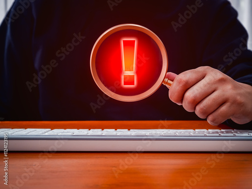 Caution alert, fraud, red exclamation mark icon, system hacked error sign, malware, attention danger symbol warning showing in magnifying glass lens, holding by person hand who working with computer. photo