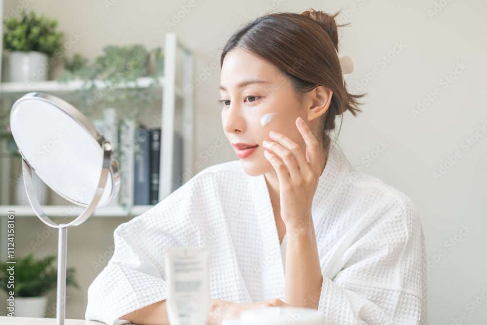 Facial beauty skin care, smile of pretty asian young woman in bathrobe looking at mirror, hand applying moisturizer lotion on her face, putting cream treatment before makeup cosmetic routine at home. Facial beauty skin care, smile of pretty asian young woman in bathrobe looking at mirror, hand applying moisturizer lotion on her face, putting cream treatment before makeup cosmetic routine at home.