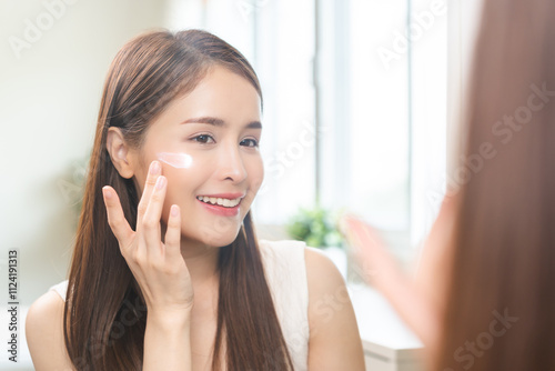 Facial beauty skin health care, smile of cute asian young woman looking at mirror, hand applying moisturizer lotion on her face, female putting cream treatment before makeup cosmetic routine at home.