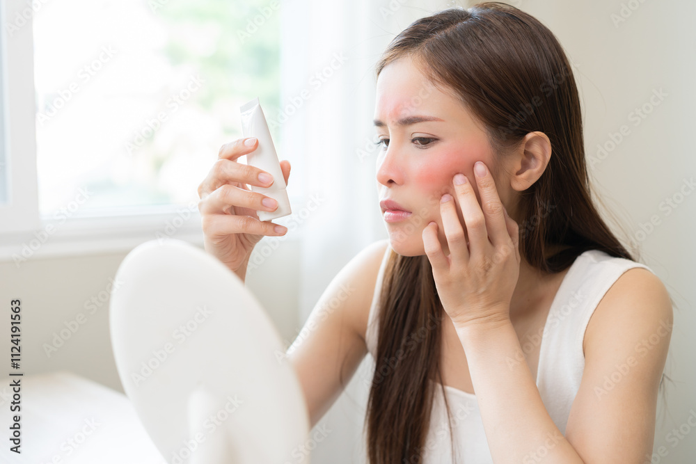 Dermatology, asian young woman looking at mirror, holding cream tube in hand, expression worry and itch, itchy allergy or allergic sensitive reaction, red or rash on face. Beauty care of skin problem Dermatology, asian young woman looking at mirror, holding cream tube in hand, expression worry and itch, itchy allergy or allergic sensitive reaction, red or rash on face. Beauty care of skin problem