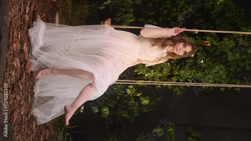 Caucasian woman with long hair in elegant white lace dress sits on wooden swing among dark foliage. Natural cosmetics or perfume concept.