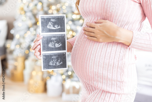 Young pregnant woman holding an ultrasound shot of her baby at home on Christmas Eve. Three photos fetus in the womb. Normal course of pregnancy. Third trimester December. successful ivf. surrogacy. 