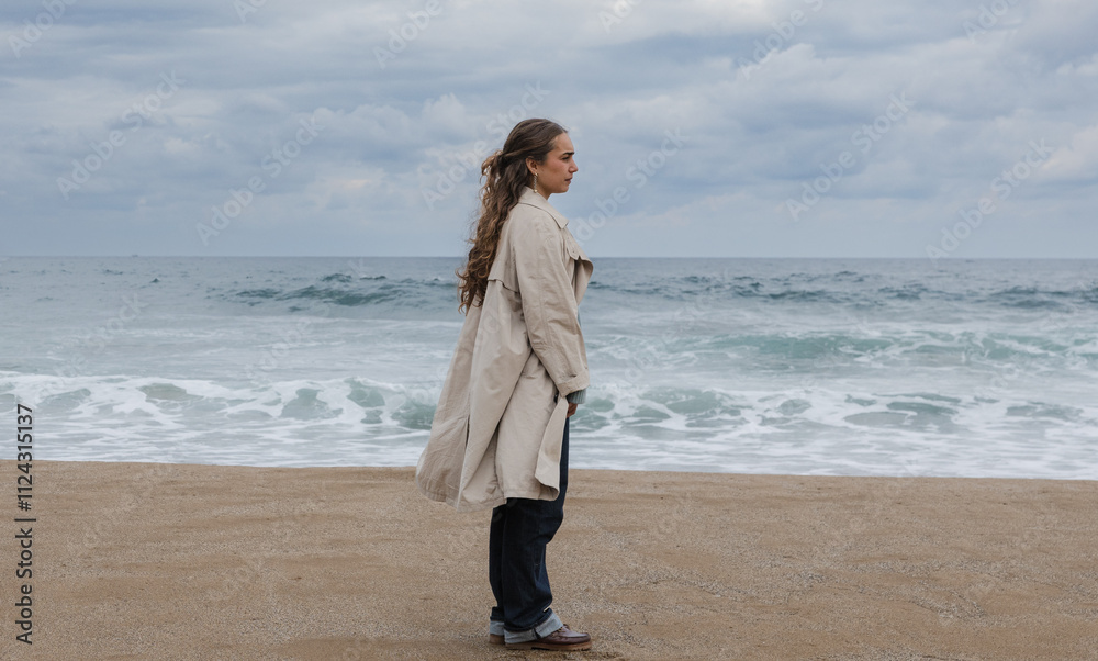 Young woman in a trench coat gazing at the ocean on a cloudy day Young woman in a trench coat gazing at the ocean on a cloudy day