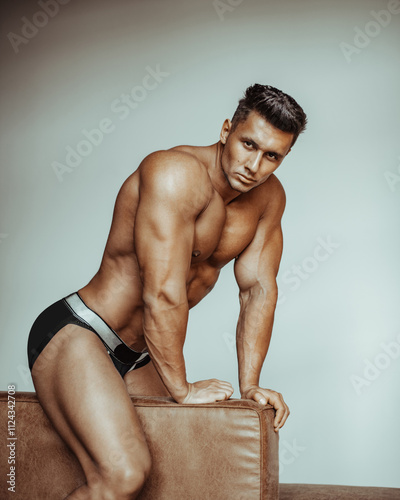 Handsome muscular man in black underwear posing on sofa. Male fitness model in briefs sitting astride sofa back. Naked brunette with six pack abs posing in studio.