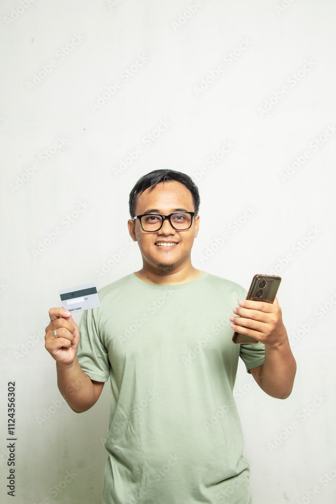 Asian man with glasses looking and smiling at camera while holding mobile phone and credit card isolated on white background Asian man with glasses looking and smiling at camera while holding mobile phone and credit card isolated on white background