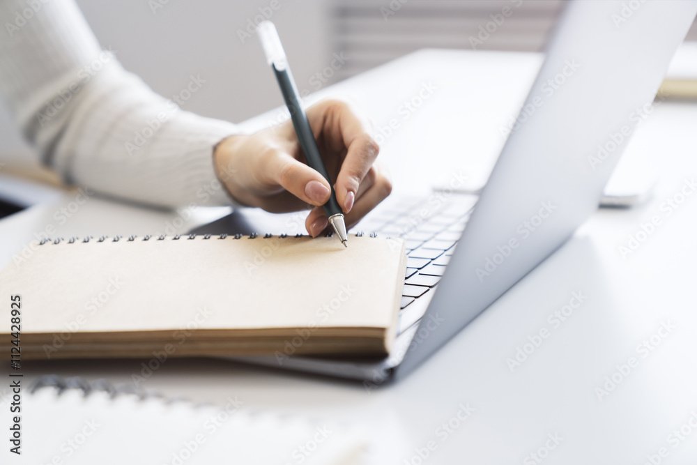 The image focuses on a woman's hand scribbling in a notepad on a stylish laptop, set against a blurry scene The image focuses on a woman's hand scribbling in a notepad on a stylish laptop, set against a blurry scene