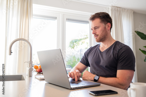 Working at home in the kitchen. caucasian male sitting at the kitchen counter behind his laptop working and communicating. Wearing simple grey t shirt. Concentrating on work.