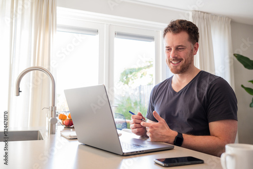 Working at home in the kitchen. caucasian male sitting at the kitchen counter behind his laptop working and communicating.Wearing simple grey t shirt. On a conference call, making gestures with hands