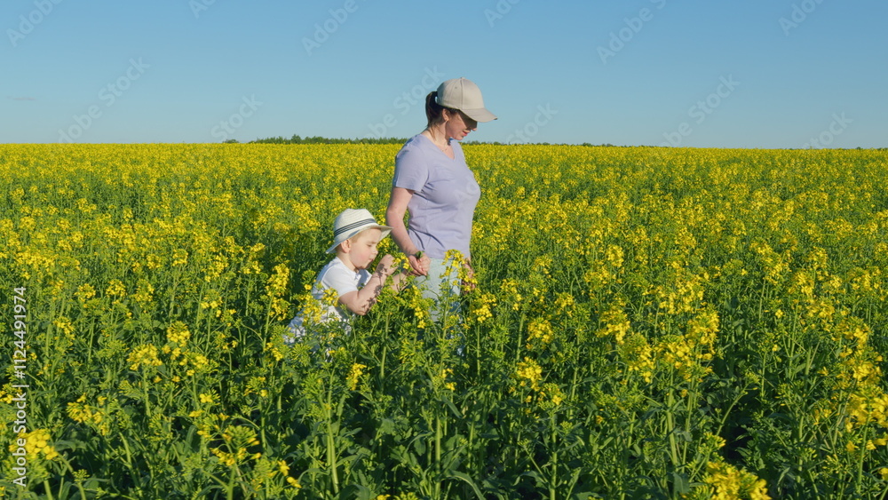 Fototapeta premium Mom Walking With Baby In Rapeseed Field. Girl With A Child Walking Along A Blooming Rapeseed. Together In Nature. Mother Day Concept.