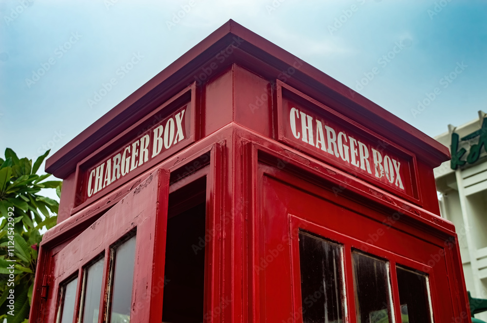 A red Charger Box booth resembling a classic British telephone booth, set against a clear sky and greenery, offering a unique and nostalgic charging station