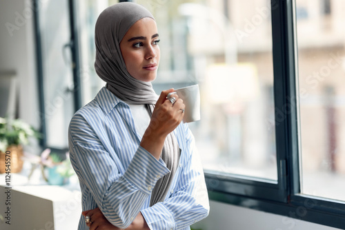 Young woman holding coffee cup and looking through window at home photo