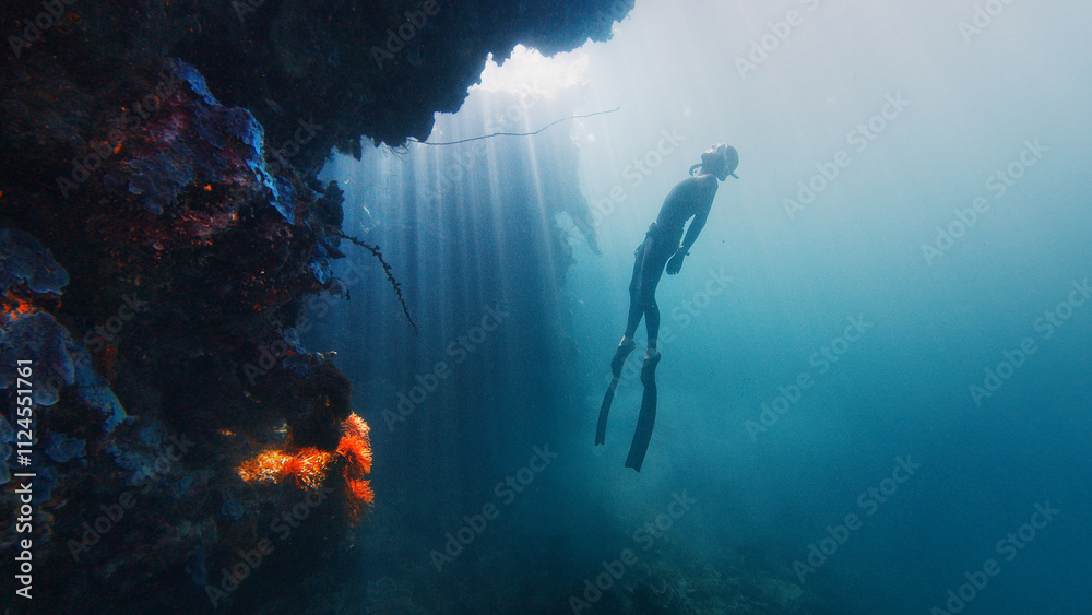 Fototapeta premium Freediver gracefully descending along a vibrant reef wall bathed in sun rays. Raja Ampat, Indonesia
