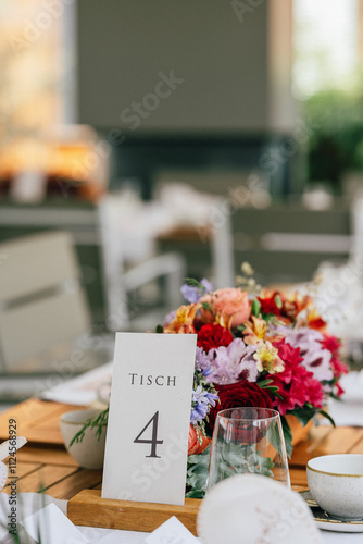 Ein festlich gedeckter Tisch mit einer Tischnummer-Karte „Tisch 4“ und einem bunten Blumenarrangement als Mittelpunkt. Die Szene wirkt modern und einladend, ideal für eine Hochzeit. photo