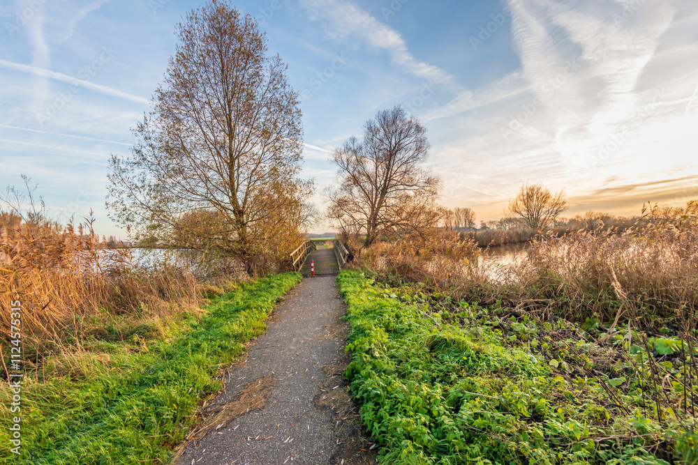Cycling and walking path with a simple wooden bridge over the water. It is at the end of a sunny and windless day in the autumn season. Most of the trees are bare and the reeds are yellowed.