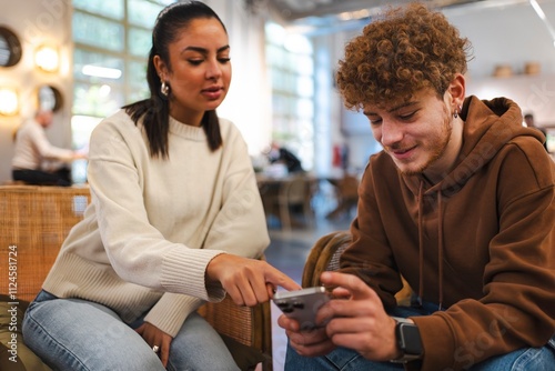 A woman in a beige sweater sitting next to a man wearing a brown hoodie indoors, pointing at his smartphone and discussing something in a casual, friendly setting photo