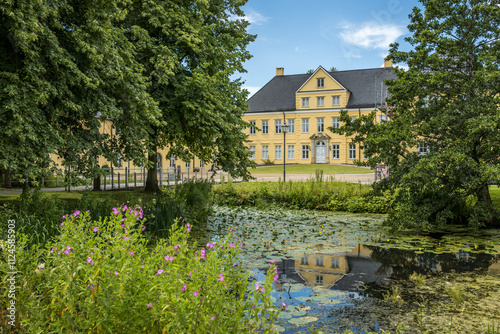 Schleswig-Holstein State Archives building in Schleswig, Germany, reflected in a tranquil pond with water lilies and surrounding greenery. photo