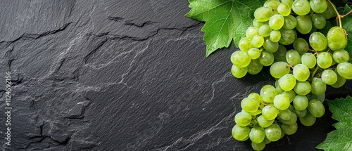 Fresh green grapes are arranged on a textured black slate surface, complemented by vibrant green leaves. photo