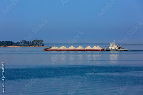 
A tugboat pulls a barge loaded with mounds of sand across a calm blue river. The serene scene includes a distant green island, blending nature with industrial activity under a clear sky.