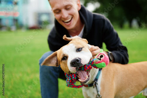 Photography Close-up of a dog holding a toy in its mouth, with a smiling owner in the background