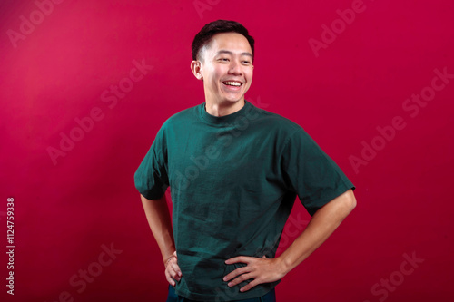 An Asian man in a casual green t-shirt strikes a confident pose with hands on hips, smiling brightly against a bold red background, exuding positivity and charm, perfect for modern marketing visuals.