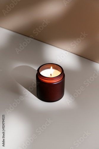 Soy wax burning candle in a dark glass jar on a beige background with shadows, minimalistic still life