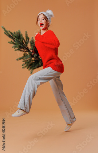 Cheerful young woman in red jumper and hat posing with christmas tree in happy running pose, isolated on orange studio background. photo