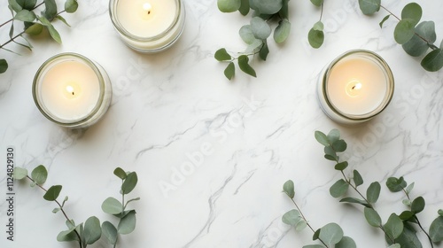 Overhead view of candles in glass holders with eucalyptus on a white marble background with space for text