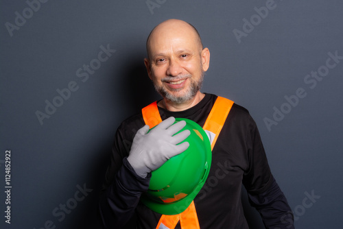 Photo of a civil worker in gloves and reflective vest holding a green helmet and looking at the camera. Isolated on gray background.