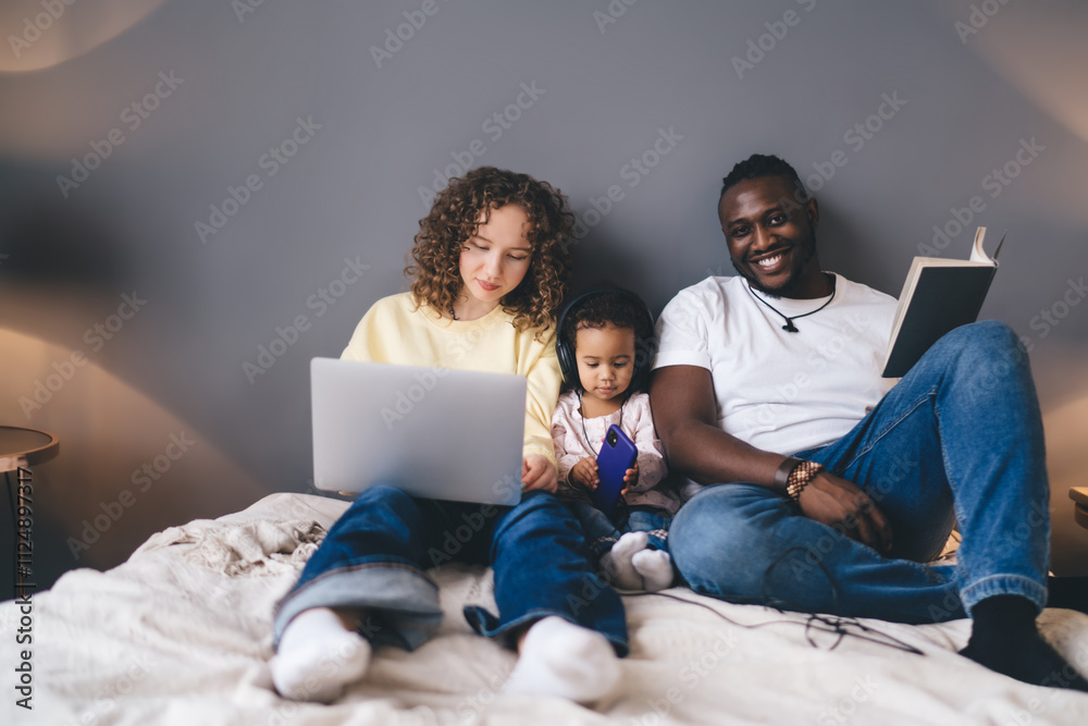 Curly-haired mother using laptop computer while father smiling at camera during relaxed moment with their young child in headphones, seamlessly integrating technology use into their cozy home interior Curly-haired mother using laptop computer while father smiling at camera during relaxed moment with their young child in headphones, seamlessly integrating technology use into their cozy home interior