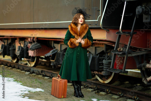 Portrait of a gorgeous red hair woman in vintage fur coat and elegant black hat with red suitcase standing near sunny retro car. Dreamy Lady traveler on a winter station. Warm art fairytale.
