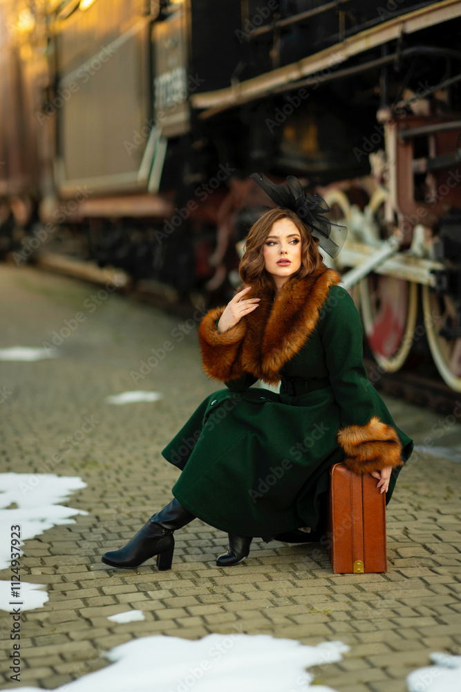 Naklejka premium Portrait of a gorgeous red hair woman in vintage fur coat and elegant black hat with red suitcase standing near sunny retro car. Dreamy Lady traveler on a winter station. Warm art fairytale. 