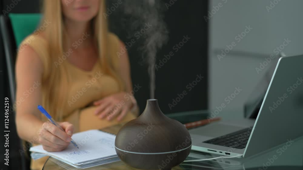Pregnant woman in her 8th-9th month working from home, sitting at a table with a laptop, discussing tasks with a colleague on a video call. A working aroma diffuser in front of her creates a calming
