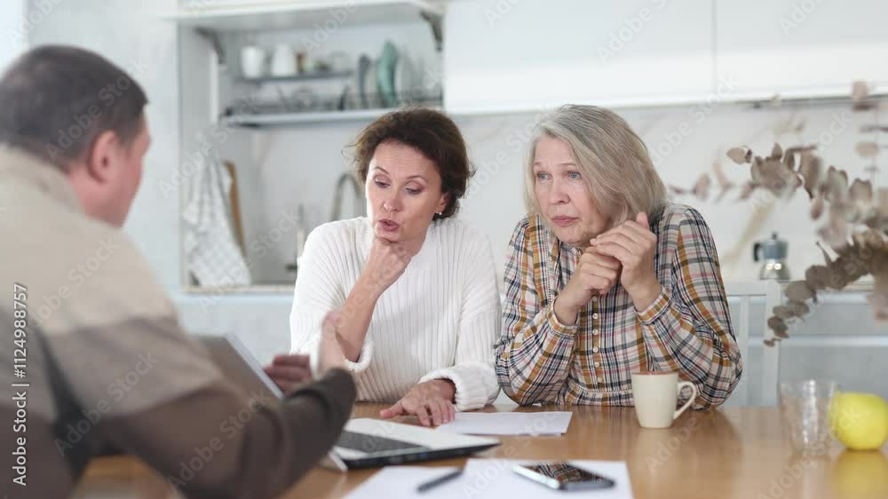 Attentive old and middle-aged women sitting in the kitchen with sheets of paper in front of them talking to male agent Attentive old and middle-aged women sitting in the kitchen with sheets of paper in front of them talking to male agent