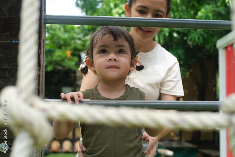 A child joyfully plays on outdoor gym equipment while their caregiver provides support in a lush, green environment. A child joyfully plays on outdoor gym equipment while their caregiver provides support in a lush, green environment.