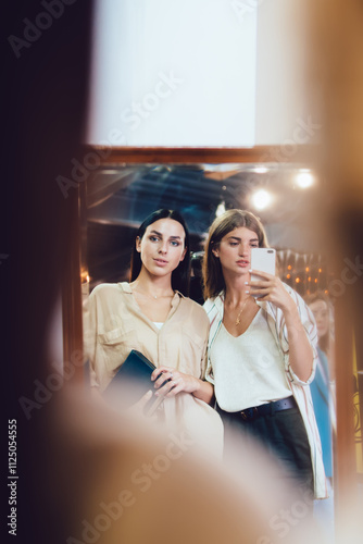 Caucasian female bloggers standing at front of mirror and photographing own reflection view using modern smartphone gadget with camera, millennial hipster girls photographing themself on leisure photo