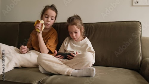 Two young sisters relax on an olive green sofa. One enjoys a banana, while the other is engrossed in a smartphone.