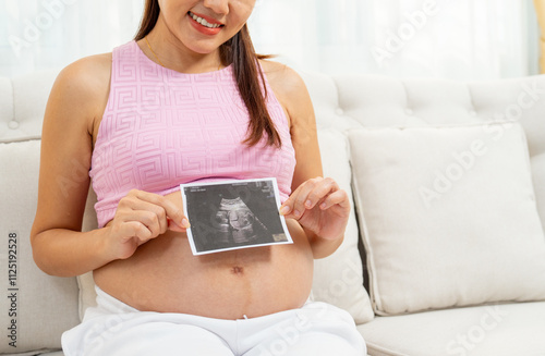 Asian pregnant woman hold with two hands ultrasound image of her baby and sit on sofa in living room and smiling.