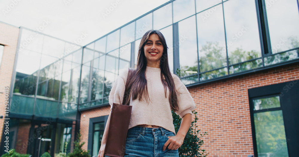 Stylish Caucasian woman posing for best view in front of modern business office with big windows. Looking at camera and smiling. Holding one hand in jeans pocket. Teaching leaving school after work.