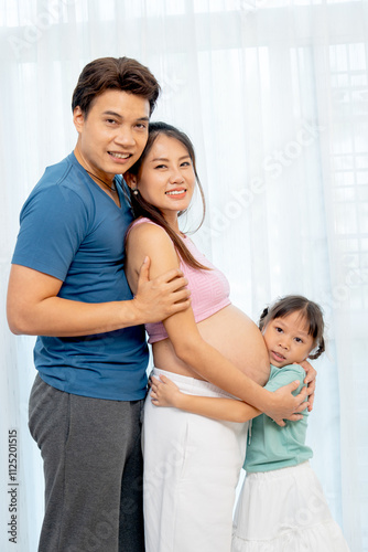 Asian family with father, pregnant mother and little girl hug together and stand in front of white curtain and look at camera.